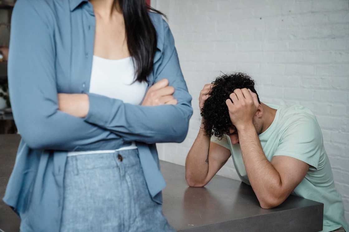 A woman stands with her arms crossed while a man sits at a table holding his head in distress.