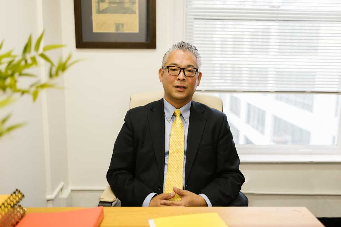 A man in a suit with glasses sits at a desk with folders and a framed document behind him.