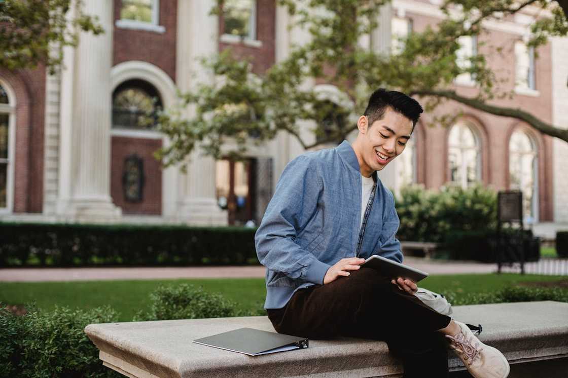 A man uses a tablet while resting on a bench in a park.