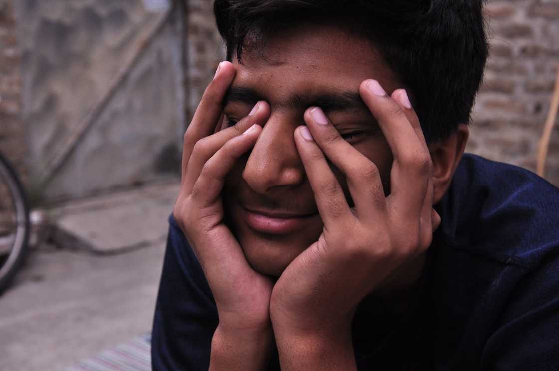 A young man sits with his face in his hands, eyes closed, appearing overwhelmed and distressed. A young man sits with his face in his hands, eyes closed, appearing overwhelmed and distressed.