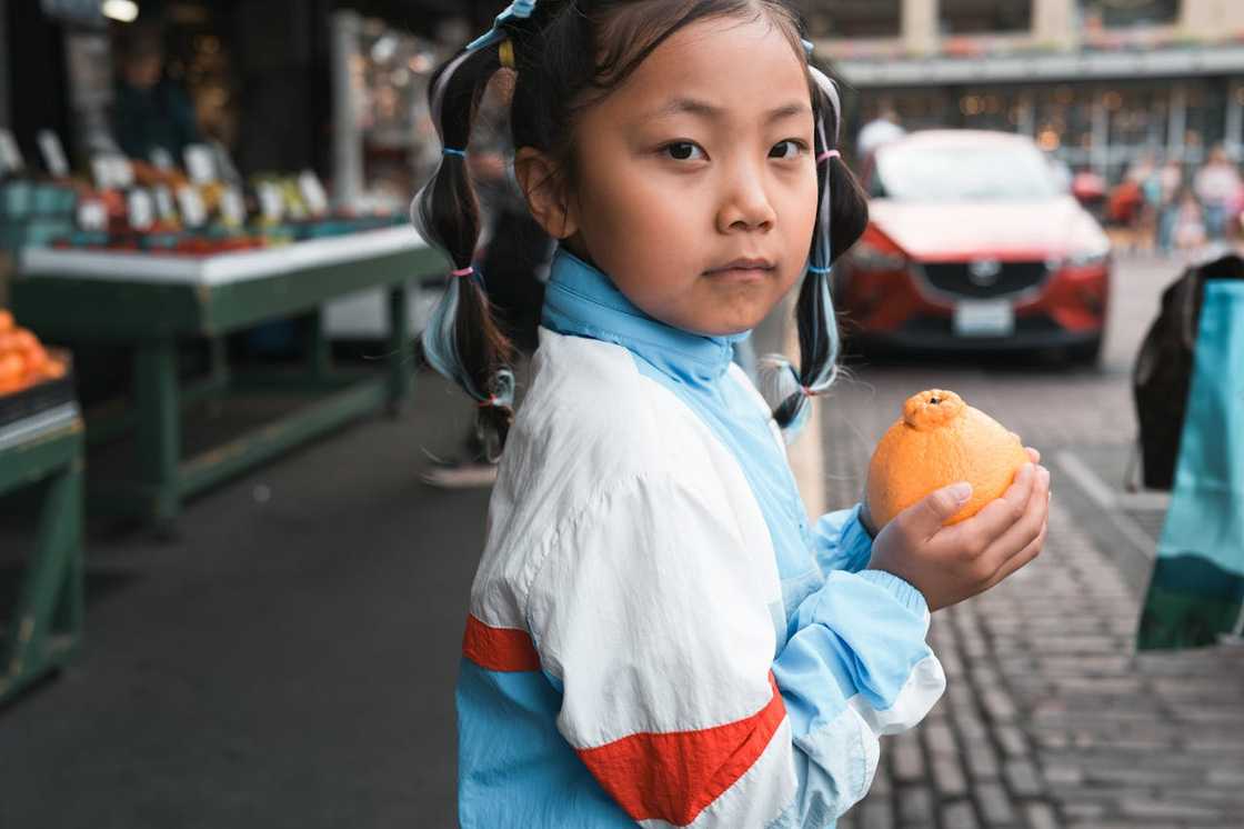 A young child stands at an outdoor market, holding an orange and looking toward the camera.