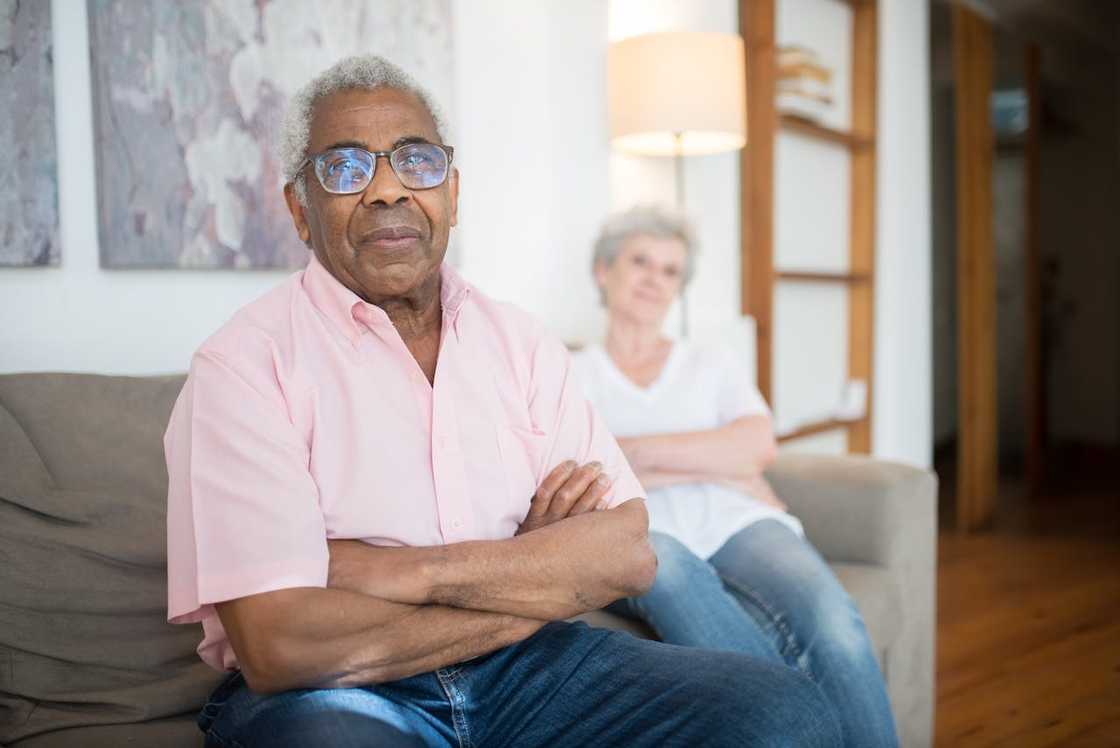 An elderly man sits on a couch with his arms crossed while a woman sits behind him.