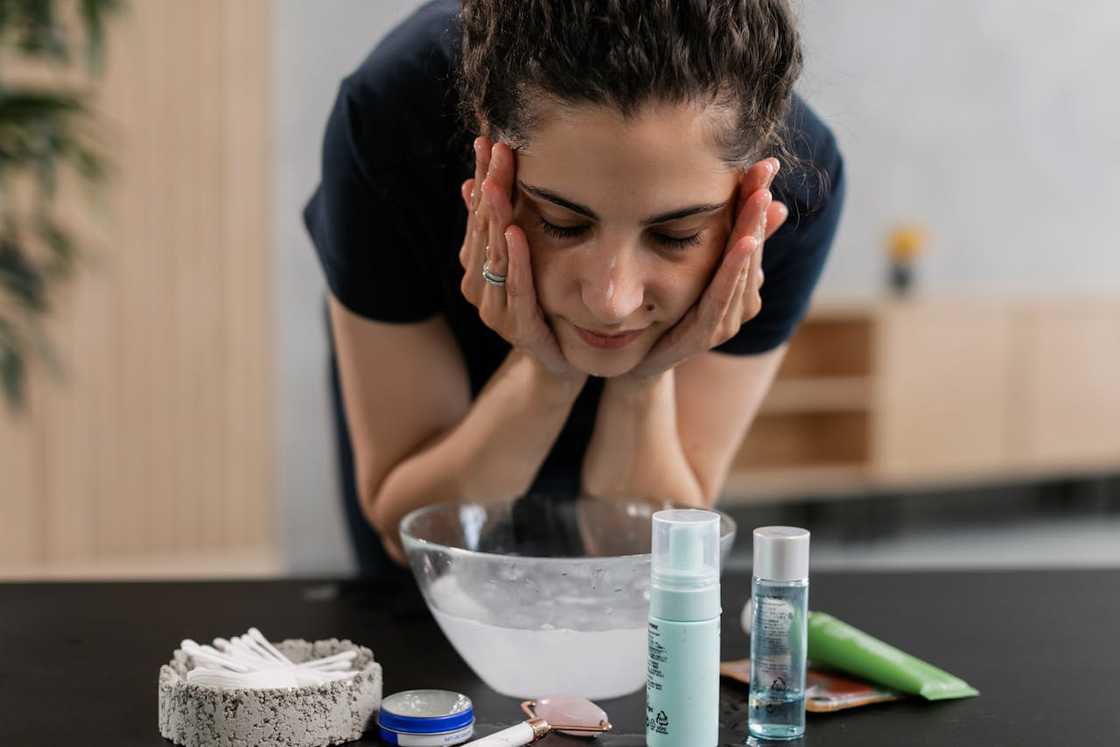 A woman washes her face over a bowl of water, with skincare products on the counter.
