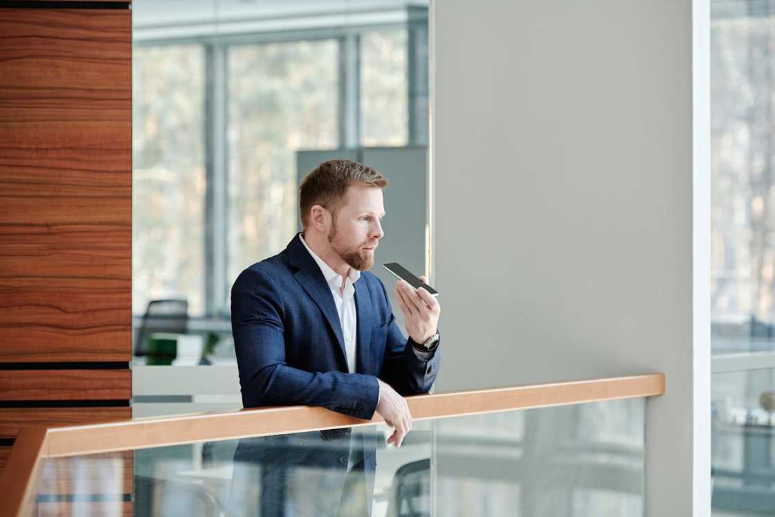 A man in a suit leans on an office railing while speaking into a smartphone. A man in a suit leans on an office railing while speaking into a smartphone.