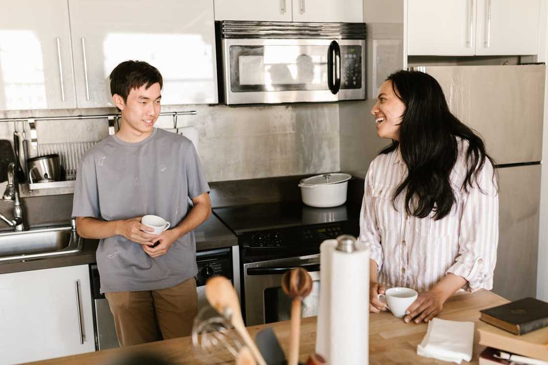 A woman and a teenage boy stand in a kitchen, holding cups and smiling at each other. A woman and a teenage boy stand in a kitchen, holding cups and smiling at each other.