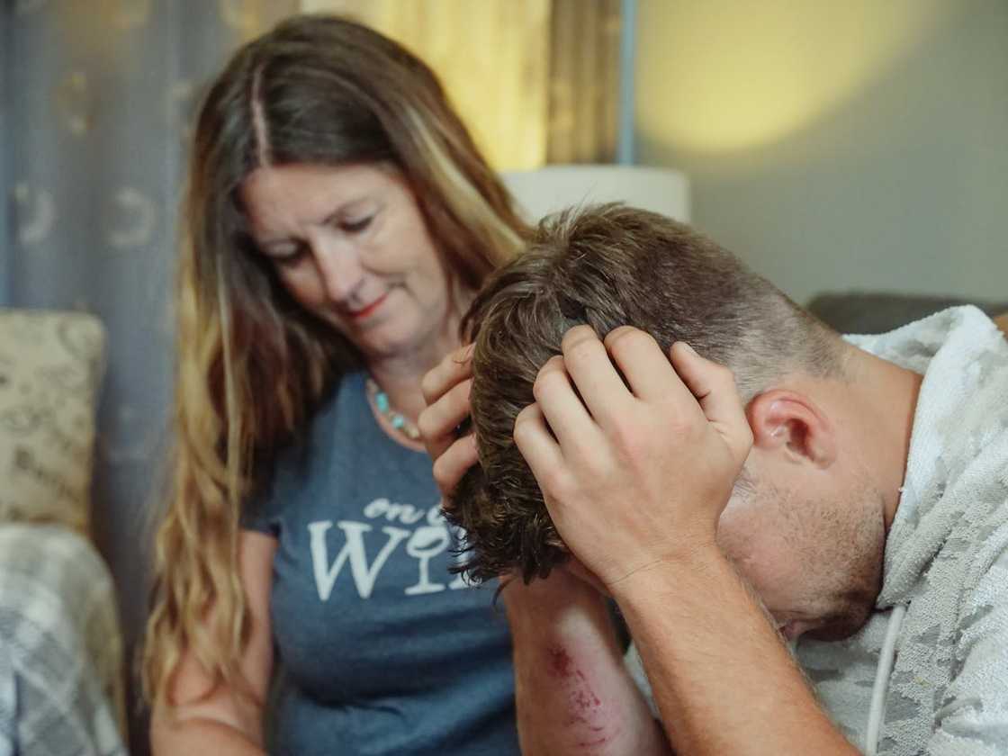 A woman sits consoles a man, appearing distressed, while she looks on supportively. A woman sits consoles a man, appearing distressed, while she looks on supportively.
