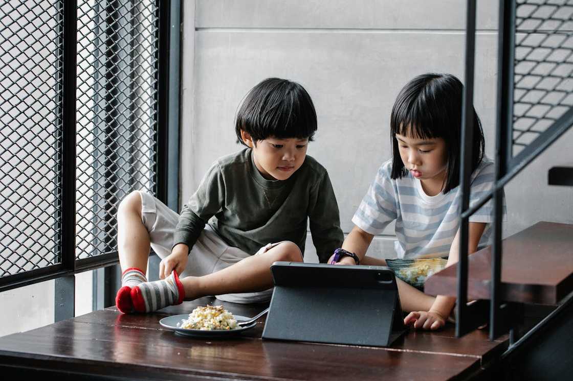 Two young children sit on a wooden floor beside a low table, watching something on a tablet together.