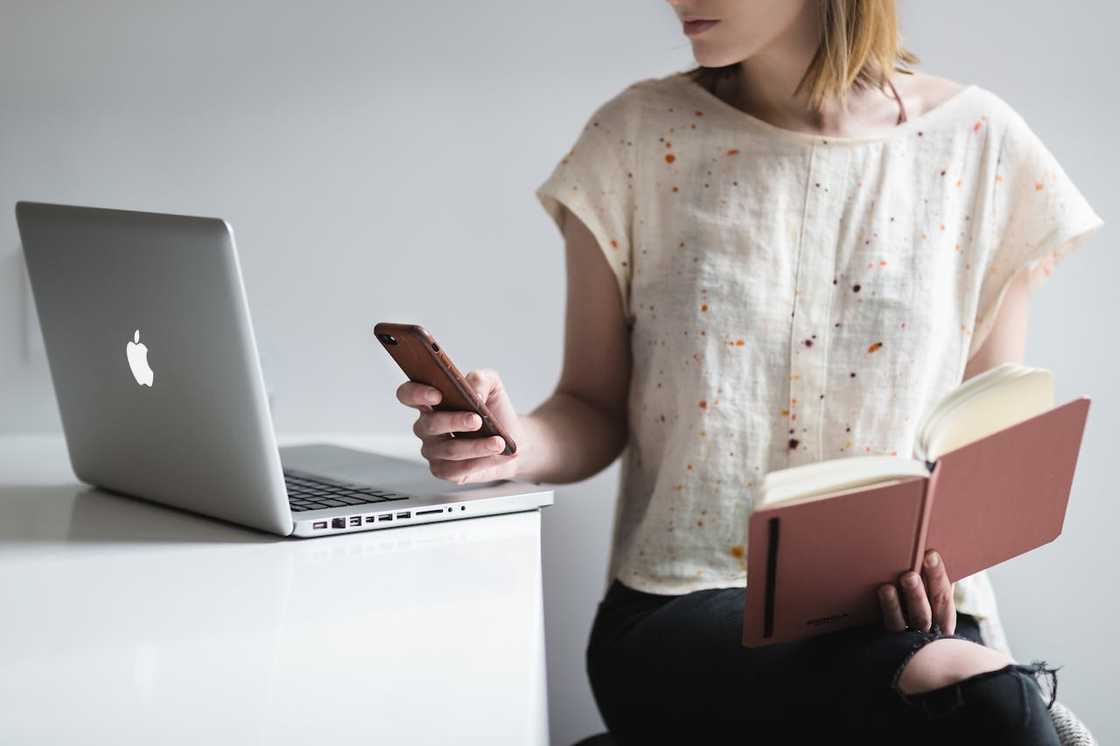 A woman sits beside a laptop, holding a phone in one hand and an open notebook in the other.