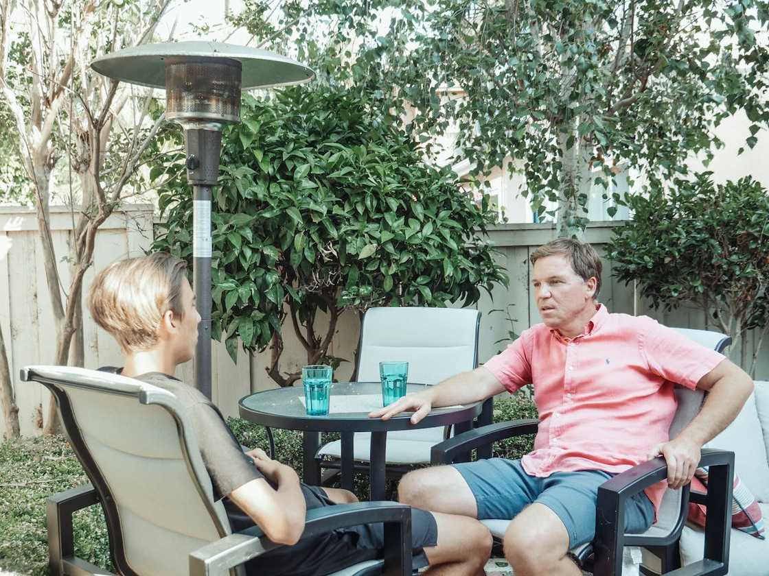 A father and teenage son sit outdoors at a patio table having a serious conversation. A father and teenage son sit outdoors at a patio table having a serious conversation.