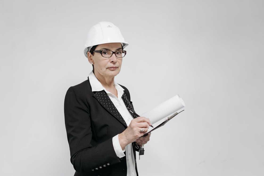 Woman wearing a hard hat and glasses, holding a clipboard and pen against a plain background.
