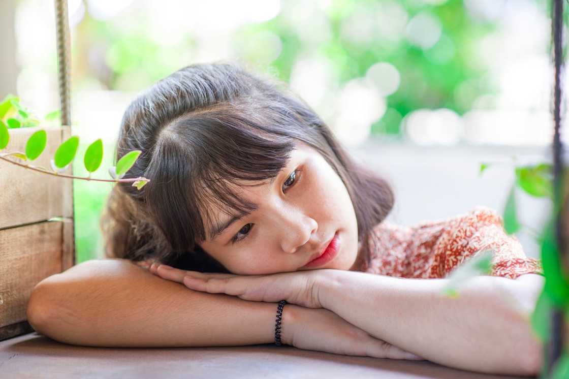 A young girl rests their head on folded arms, surrounded by soft greenery and natural light. A young girl rests their head on folded arms, surrounded by soft greenery and natural light.