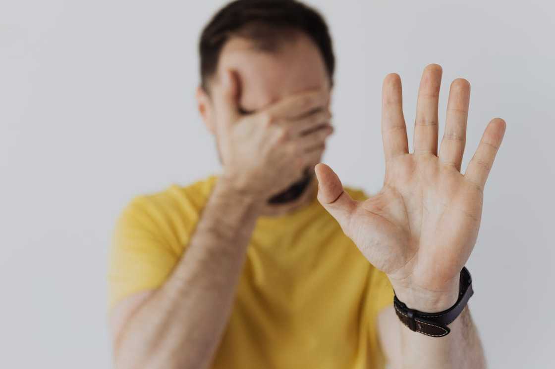 A man covers his face with one hand while raising the other palm forward in a stop or refusal gesture. A man covers his face with one hand while raising the other palm forward in a stop or refusal gesture.