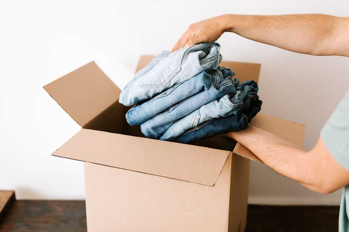 Hands placing folded clothes into a cardboard moving box.