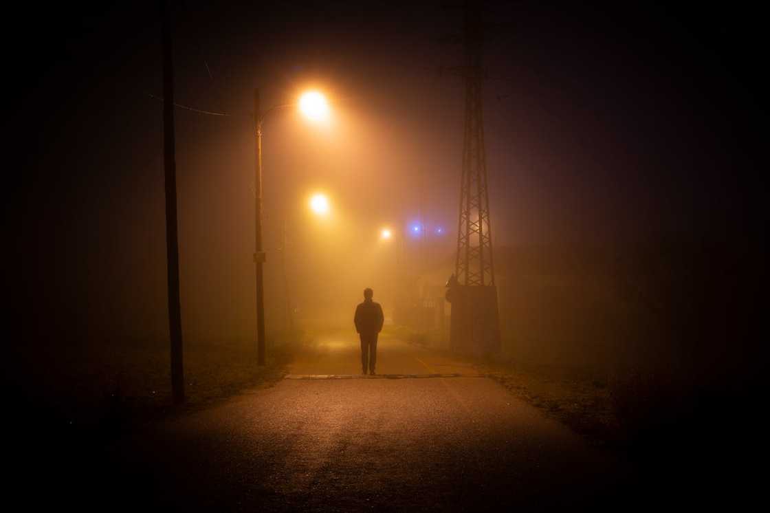 A person walks alone down a foggy street at night under streetlights.