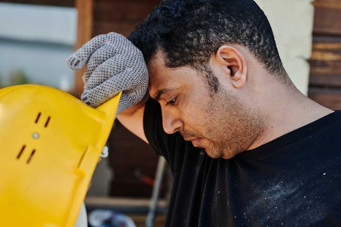 A tired construction worker wipes sweat from his forehead while holding a yellow hard hat.