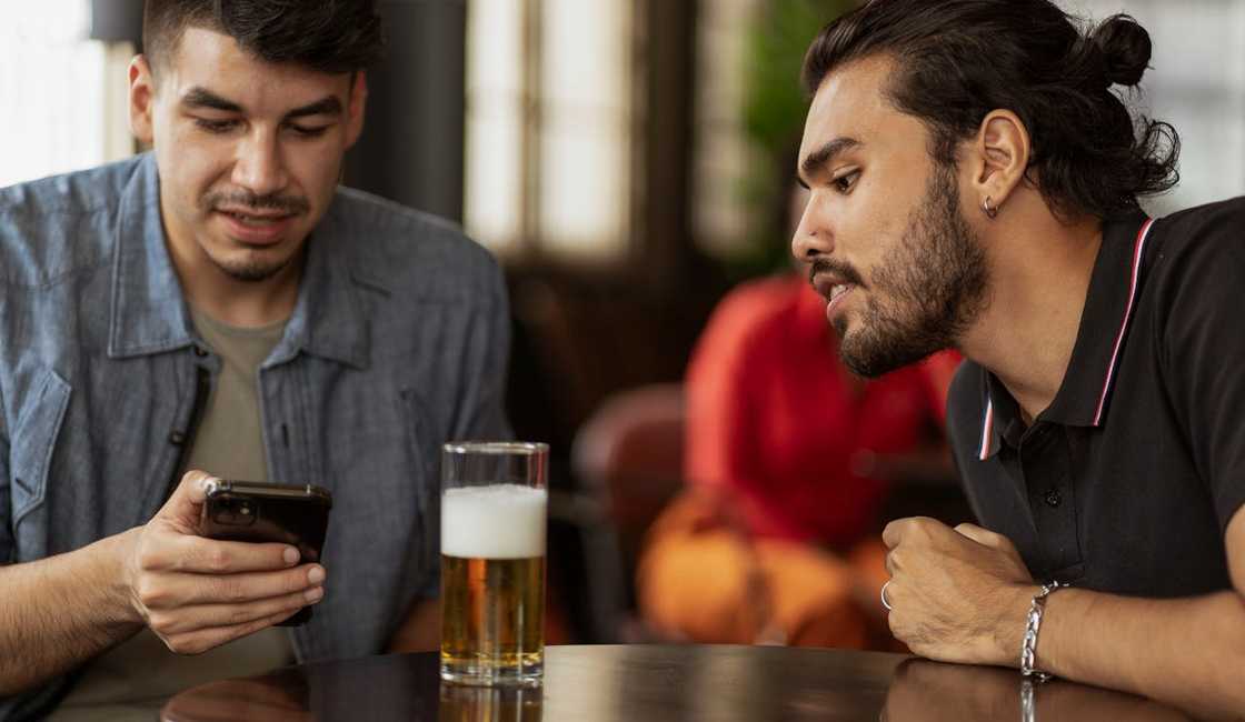 Two men sit at a table, leaning in to look at a smartphone beside a glass of beer. Two men sit at a table, leaning in to look at a smartphone beside a glass of beer.