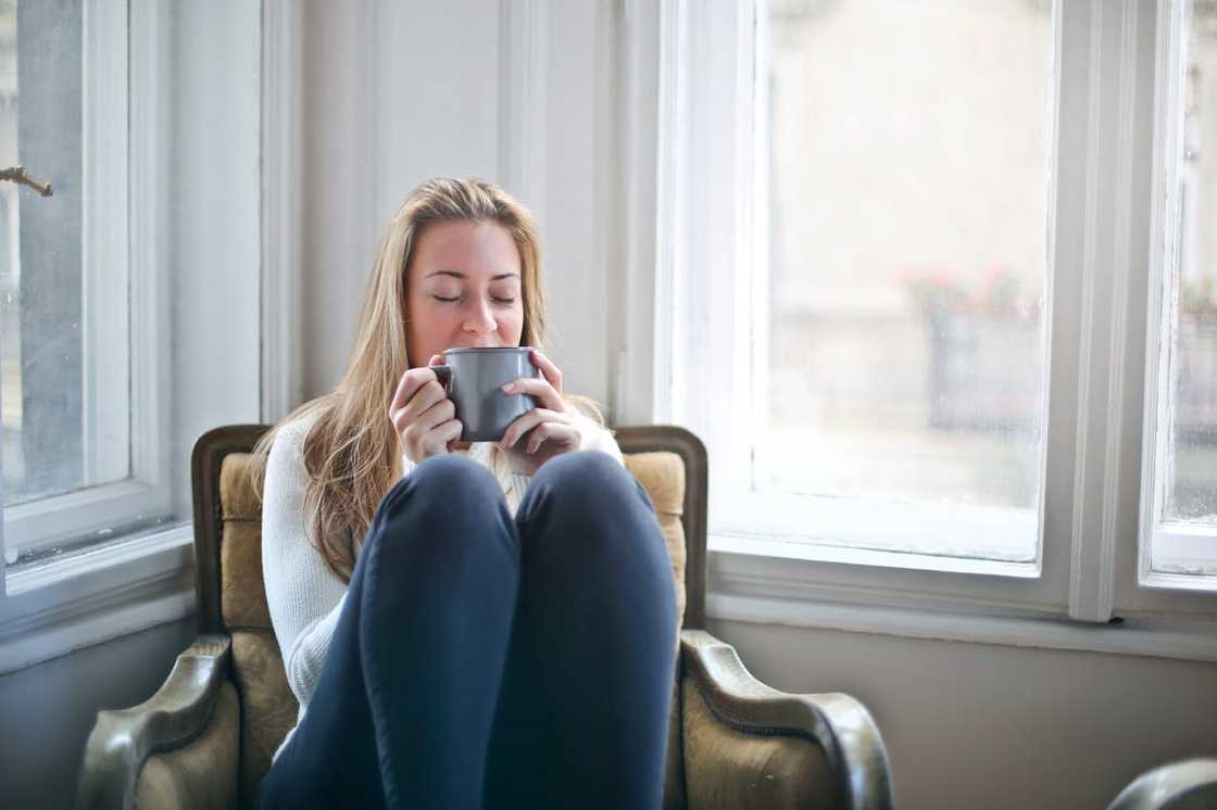 A woman sits curled up in an armchair by a window, holding a mug and relaxing quietly.