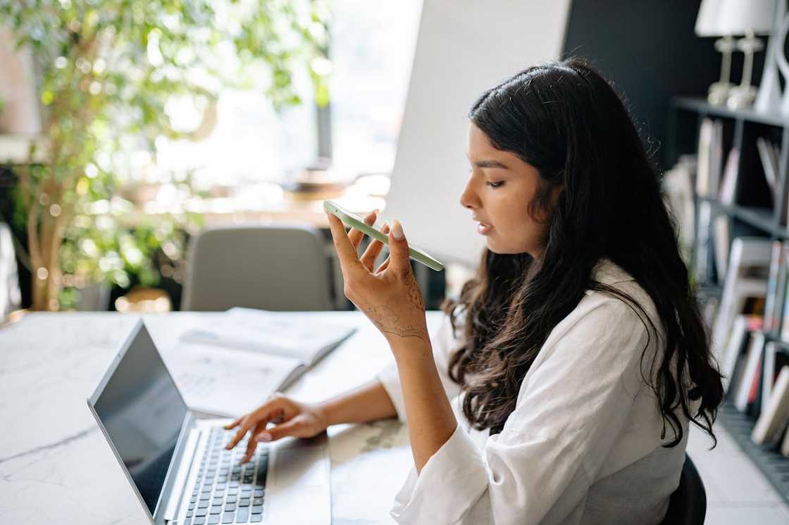 A woman speaking into her smartphone while working on a laptop at a desk. A woman speaking into her smartphone while working on a laptop at a desk.
