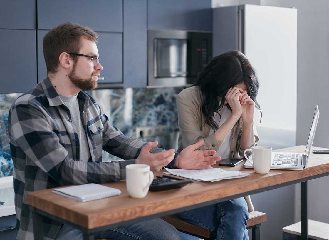 A man gestures while a distressed woman covers her face at a kitchen table with bills and a laptop.