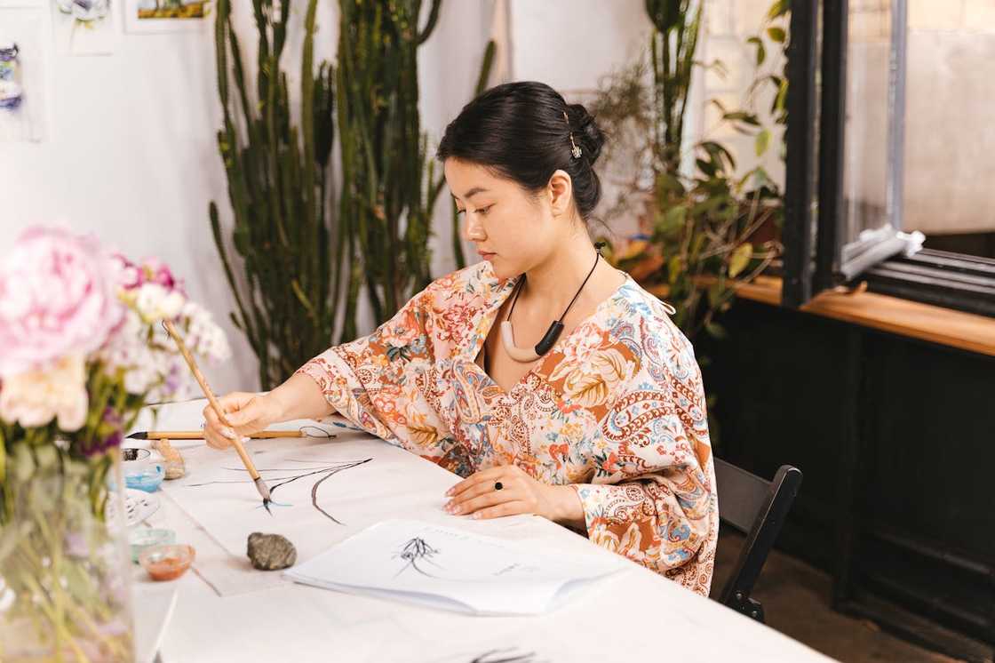 A focused artist works on a delicate brush painting in a bright, plant-filled studio.