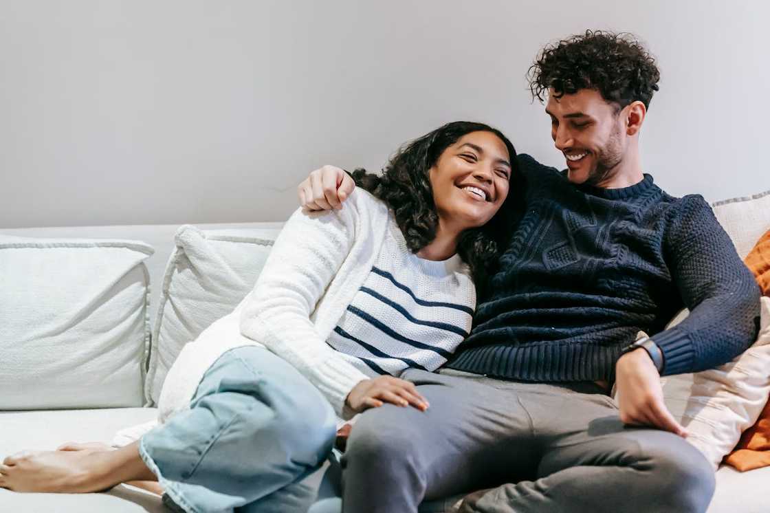 A couple sits close together on a sofa, smiling and relaxed at home.