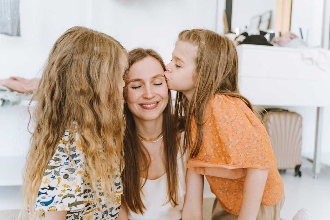 Two young girls kiss their smiling mother on the cheeks indoors.