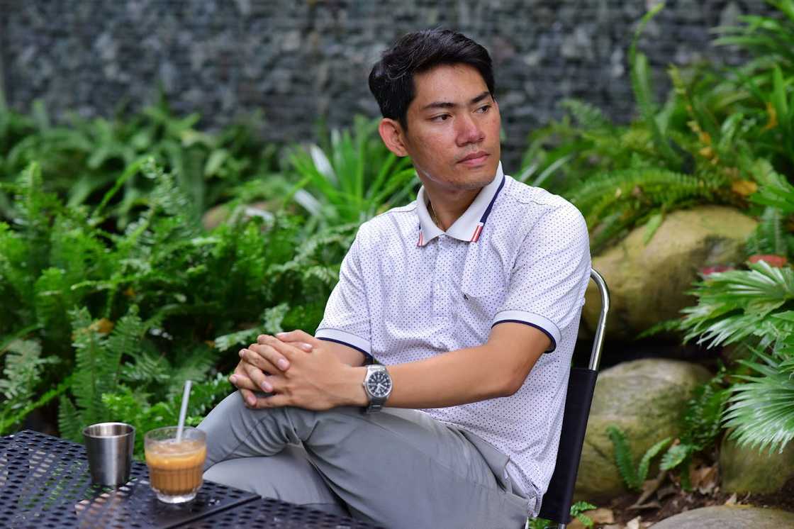 A person in a white polo shirt and grey pants sits at a metal table in a lush garden. A person in a white polo shirt and grey pants sits at a metal table in a lush garden.