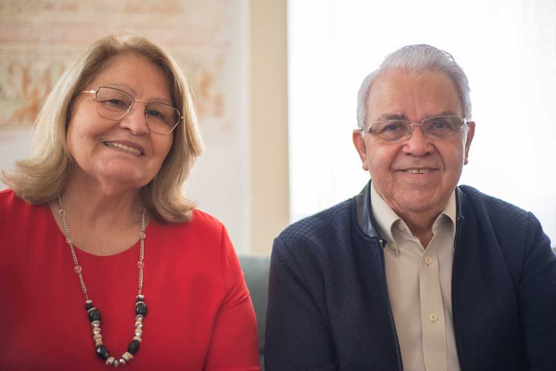 An older woman and an older man sit side by side indoors, smiling at the camera.
