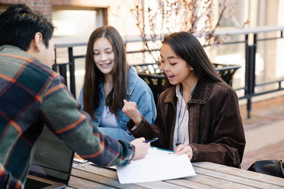 Three people sit at an outdoor table, discussing notes together. Three people sit at an outdoor table, discussing notes together.