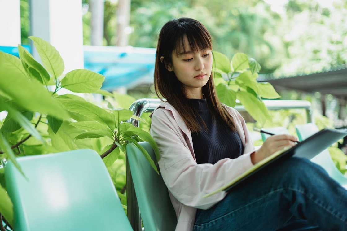 A teenager sits outdoors writing in a notebook. A teenager sits outdoors writing in a notebook.