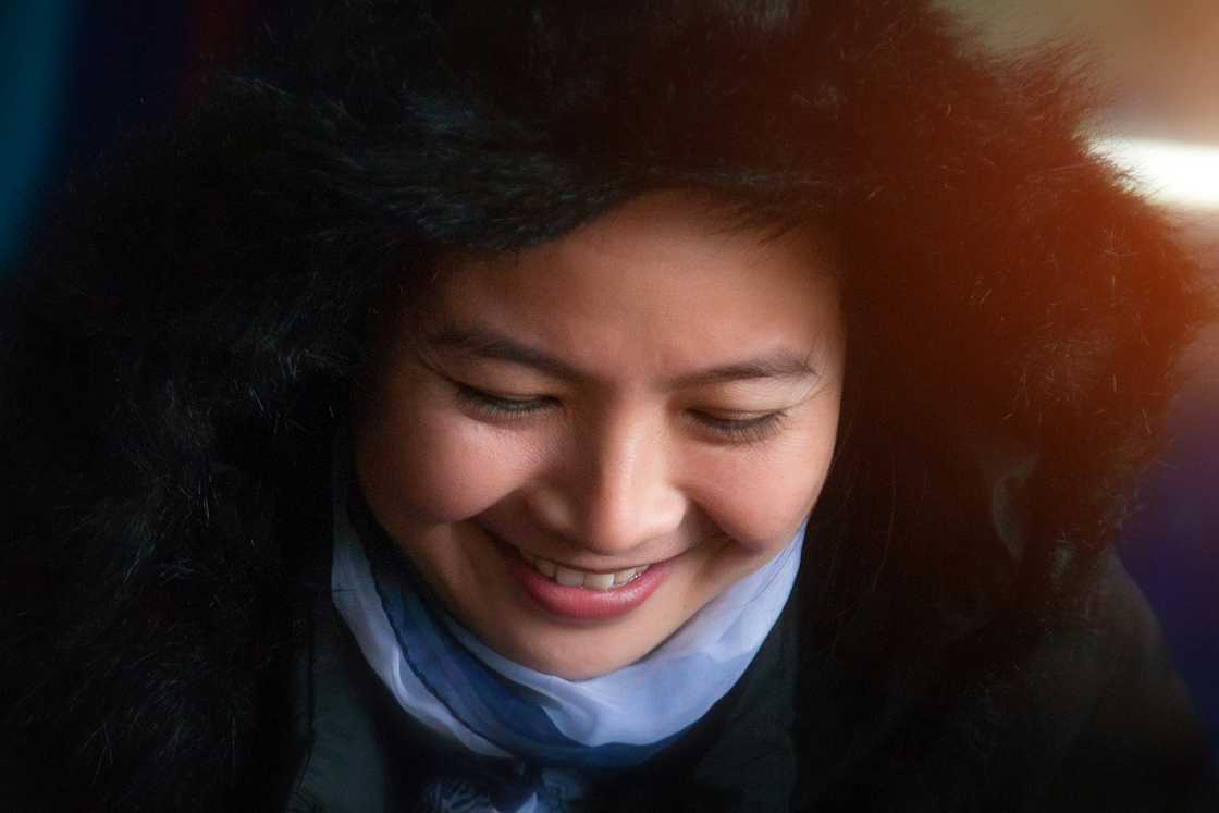 A person in a fur-lined hood and layered scarf looks down with closed eyes and a soft smile.