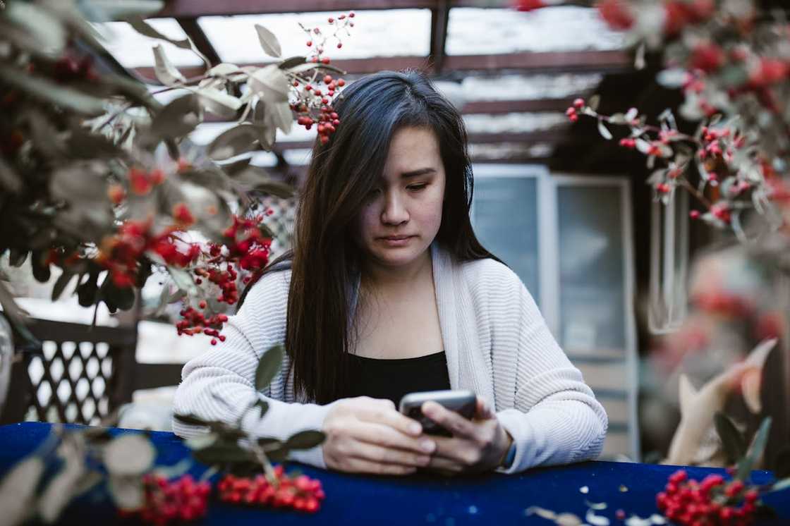 A woman sits at an outdoor table, holding a phone and looking down thoughtfully.