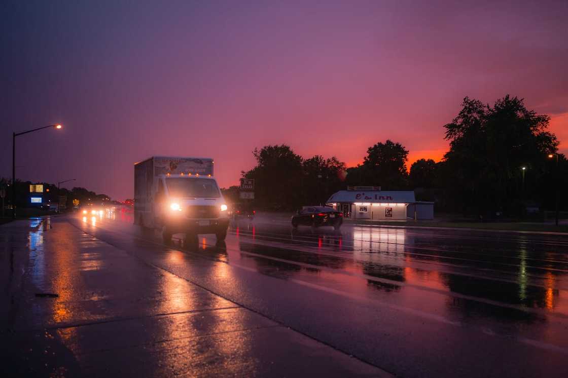 A wet road at dusk shows cars driving under streetlights after rain. A wet road at dusk shows cars driving under streetlights after rain.