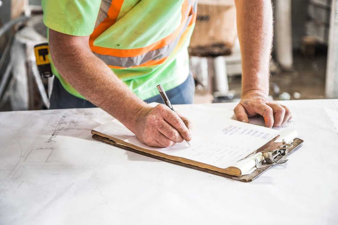 A construction worker writes notes on a clipboard over blueprints. A construction worker writes notes on a clipboard over blueprints.