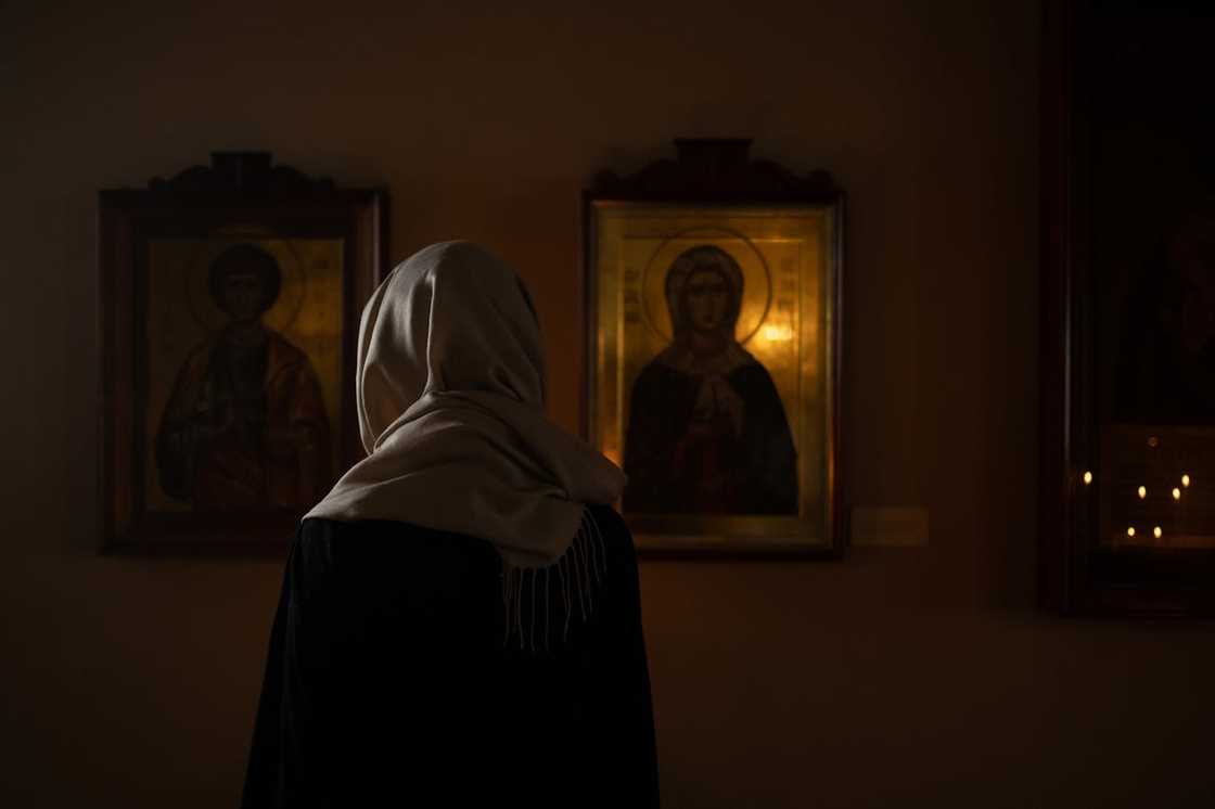 A woman wearing a headscarf stands in a dimly lit chapel, facing religious icons.