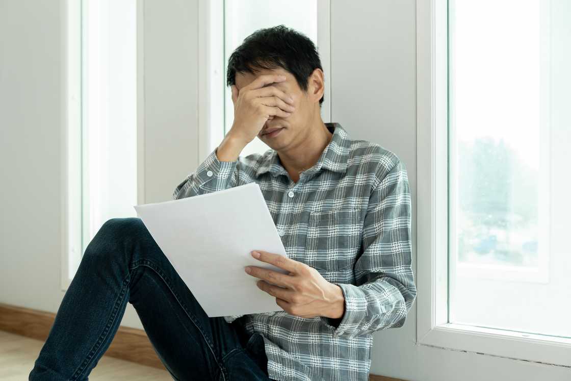 A stressed man sitting alone on the floor with a document in his hand