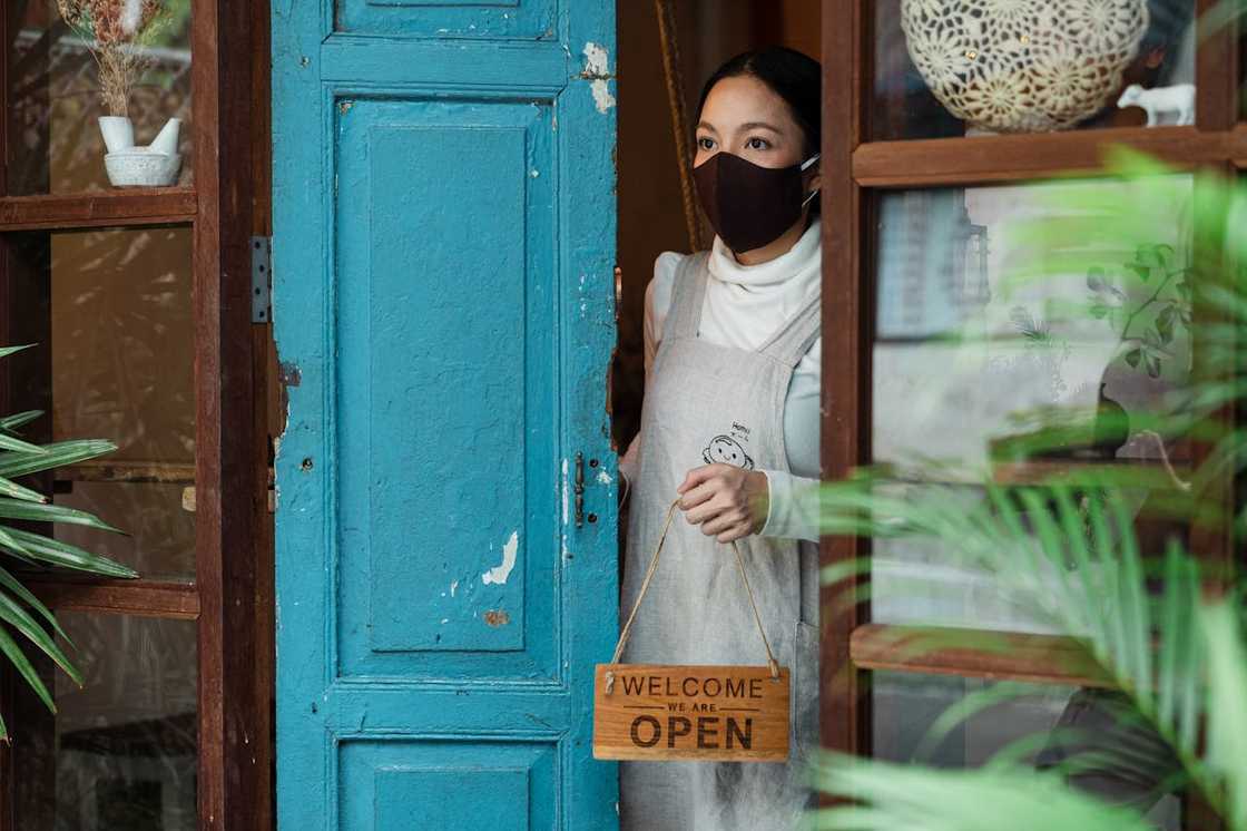 A woman wearing a face mask stands in a doorway holding a sign that says the shop is open. A woman wearing a face mask stands in a doorway holding a sign that says the shop is open.