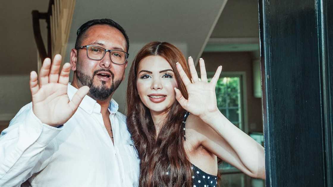 A man and woman stand in a doorway smiling and waving toward the camera. A man and woman stand in a doorway smiling and waving toward the camera.