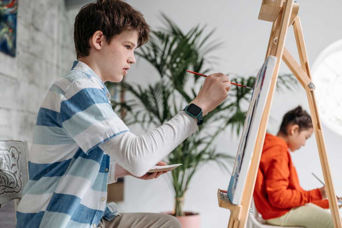 A boy paints at an easel while another child paints in the background.