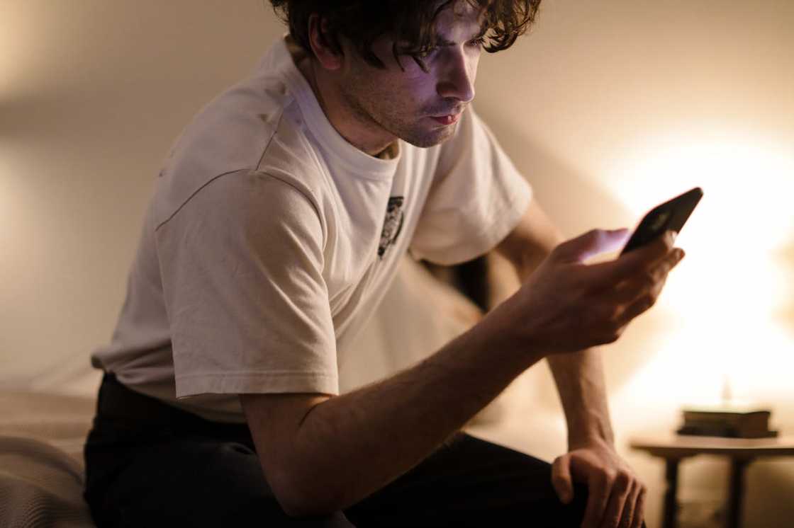 A man sits on a bed at night, leaning forward and looking at his phone. A man sits on a bed at night, leaning forward and looking at his phone.