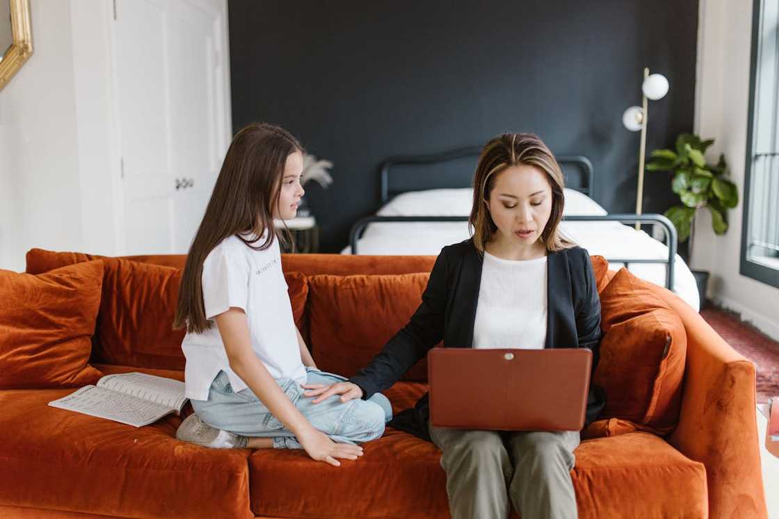 A mother is on her laptop while her daughter sits silently nearby. A mother is on her laptop while her daughter sits silently nearby.
