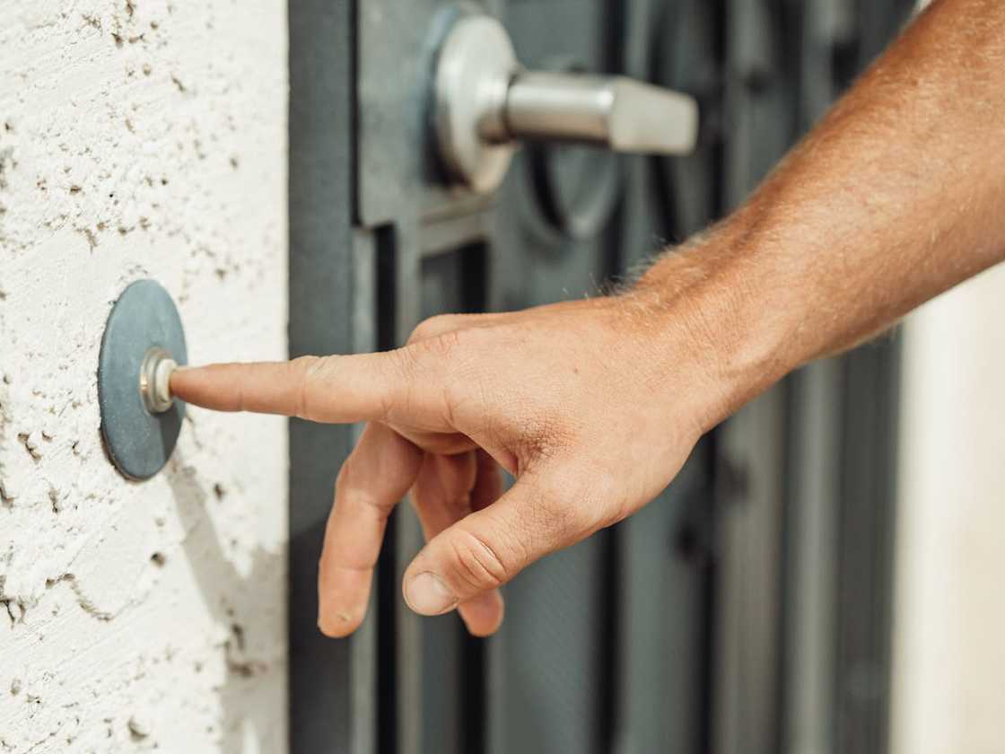 A hand presses a doorbell beside a metal gate. A hand presses a doorbell beside a metal gate.