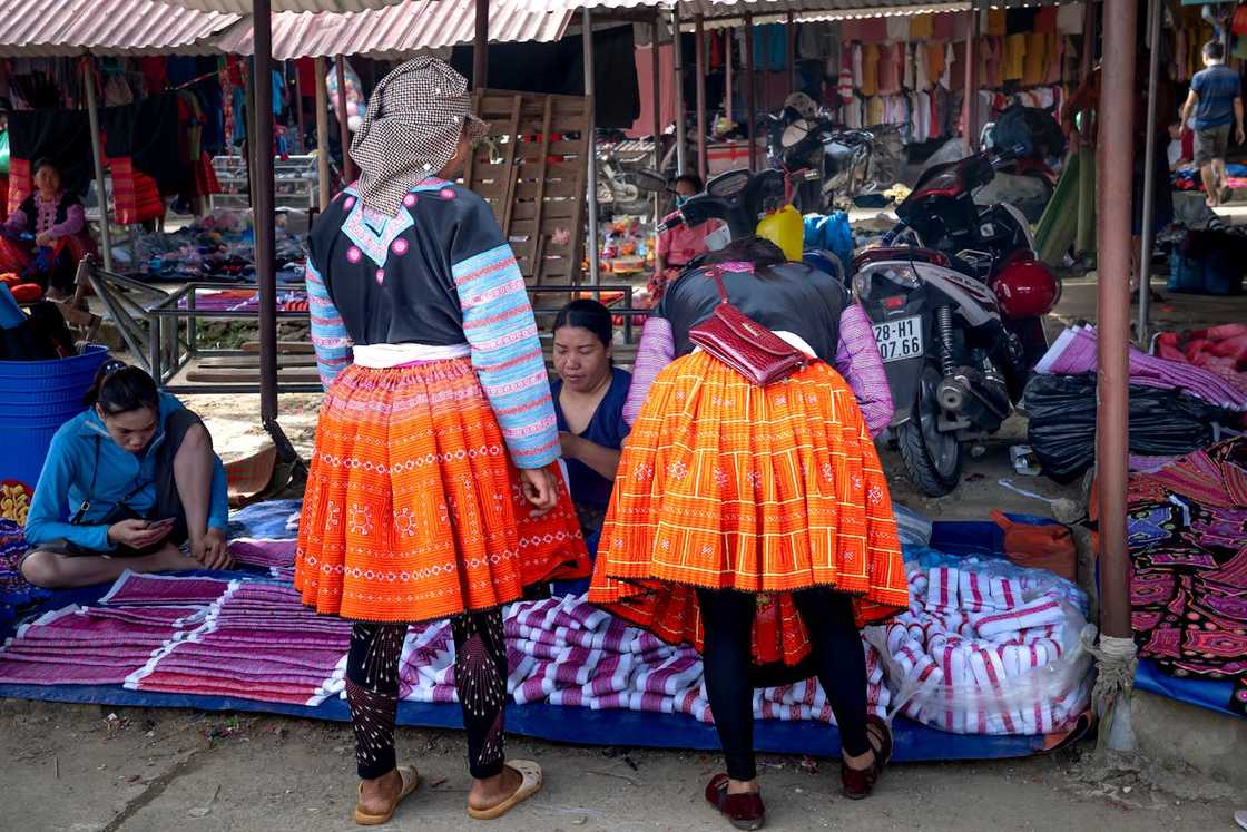 Two women in traditional dress browse textiles at an open-air market.