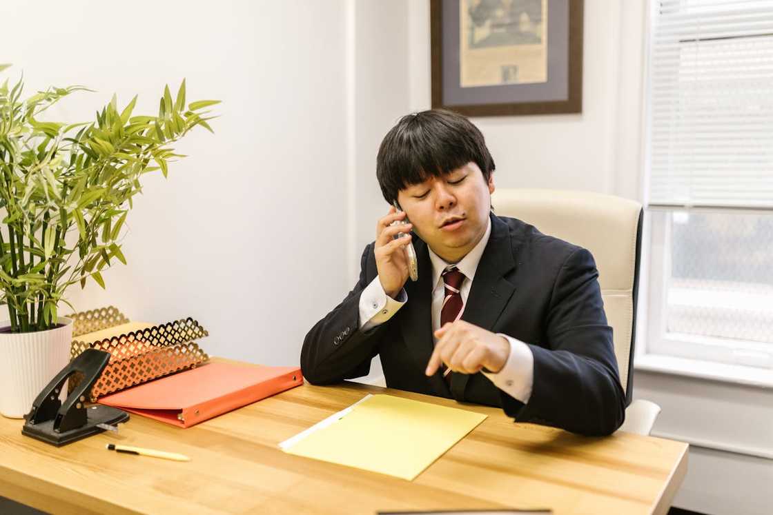 A man in a suit talks on a phone and points at a document on a desk. A man in a suit talks on a phone and points at a document on a desk.
