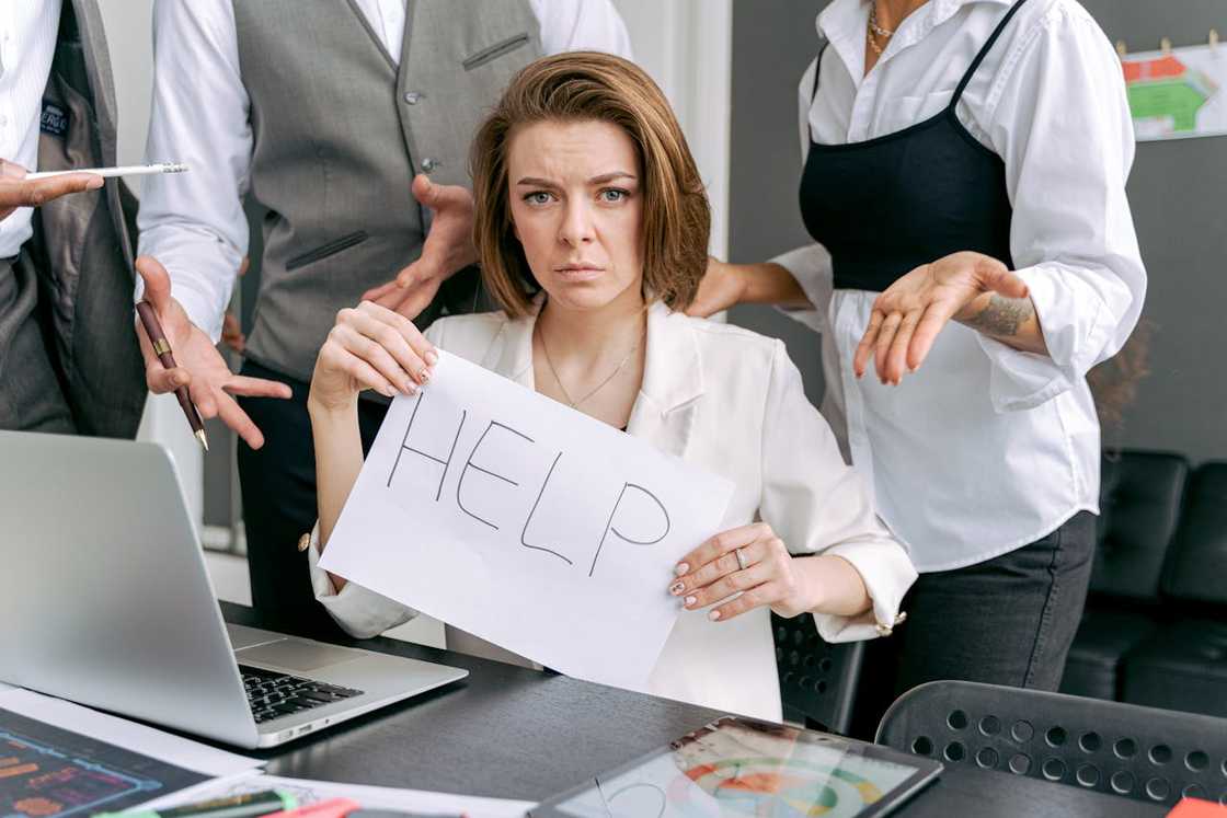 A woman holding a sign that reads “HELP” while coworkers gesture around her at a desk. A woman holding a sign that reads “HELP” while coworkers gesture around her at a desk.