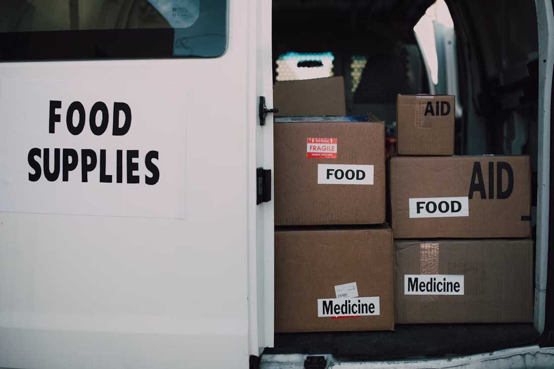 A van with its rear doors open shows stacked boxes labelled food, aid, and medicine inside. A van with its rear doors open shows stacked boxes labelled food, aid, and medicine inside.