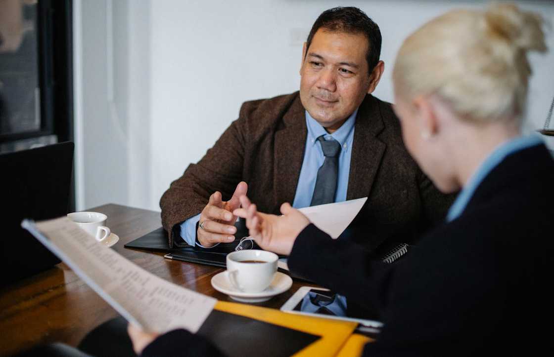 Two people sit at a table in a formal setting, discussing documents with coffee and papers spread out.