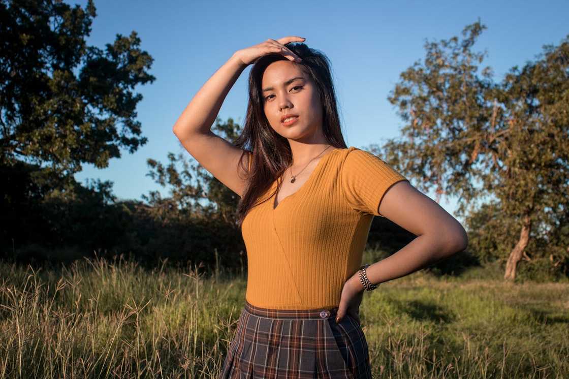 A woman standing outdoors in a grassy field, posing confidently with one hand on her head.