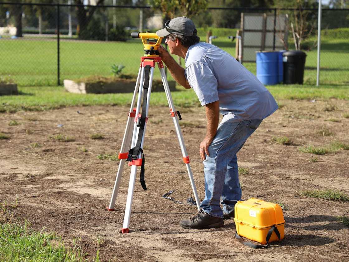 A land surveyor using a tripod-mounted level to measure ground outdoors.