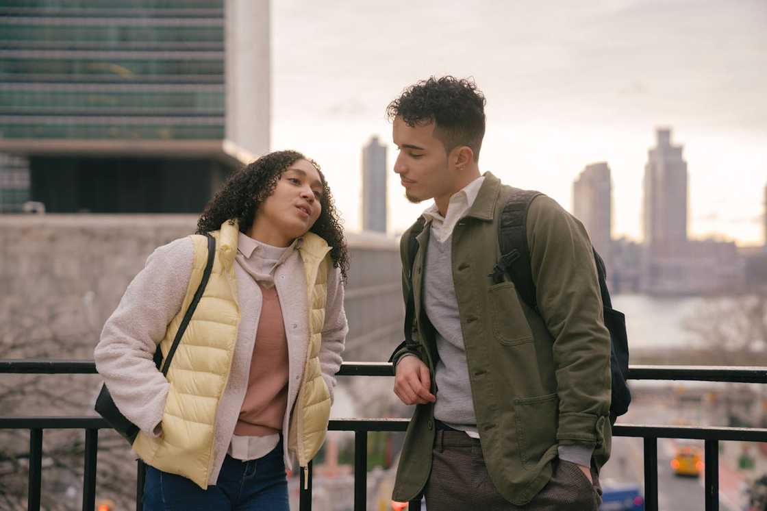 A young woman speaks seriously to a man as they stand by a railing with a city skyline behind them.